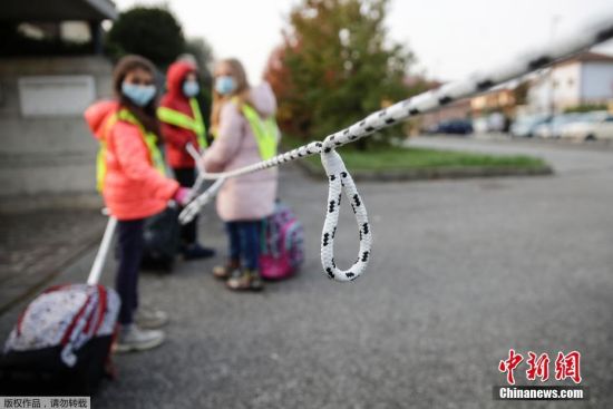 当地时间10月20日，在意大利北部贝卢斯科，孩子们拿着绳子保持社交距离，在志愿者的护送下走向学校。图为绳子上每隔一定距离就打一个结，帮助孩童确定距离。